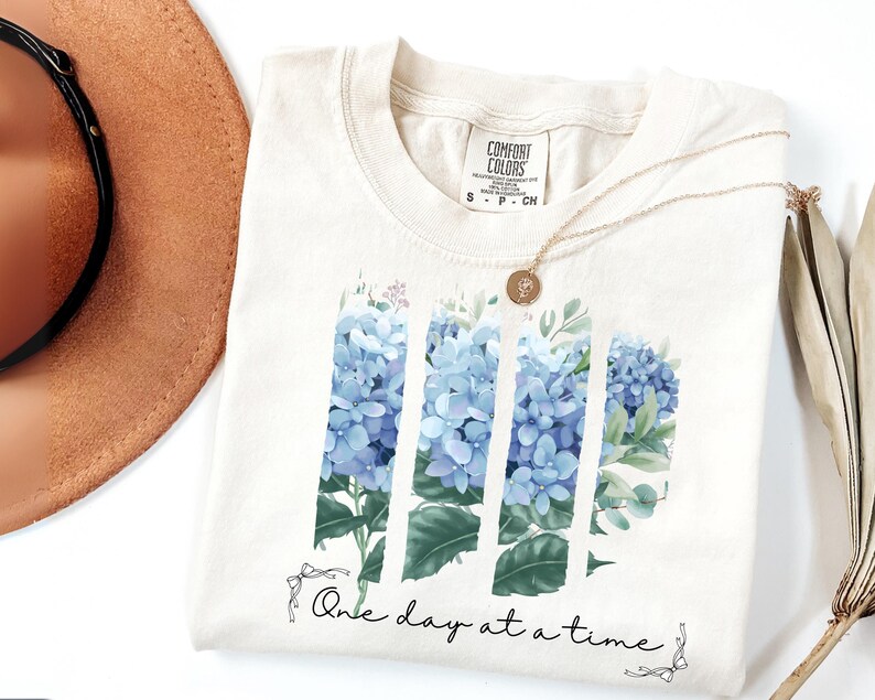 May include: A cream-colored t-shirt featuring a graphic of blue hydrangea flowers arranged in three vertical panels. Below the flowers, the text "One day at a time" is printed in a black script font, accented by small line-drawn bows on each side.