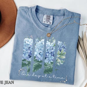 May include: A blue jean colored t-shirt featuring a graphic of four vertical panels filled with light blue hydrangea flowers and green leaves. Below the floral design, the text reads One day at a time in white cursive script with small bow accents.