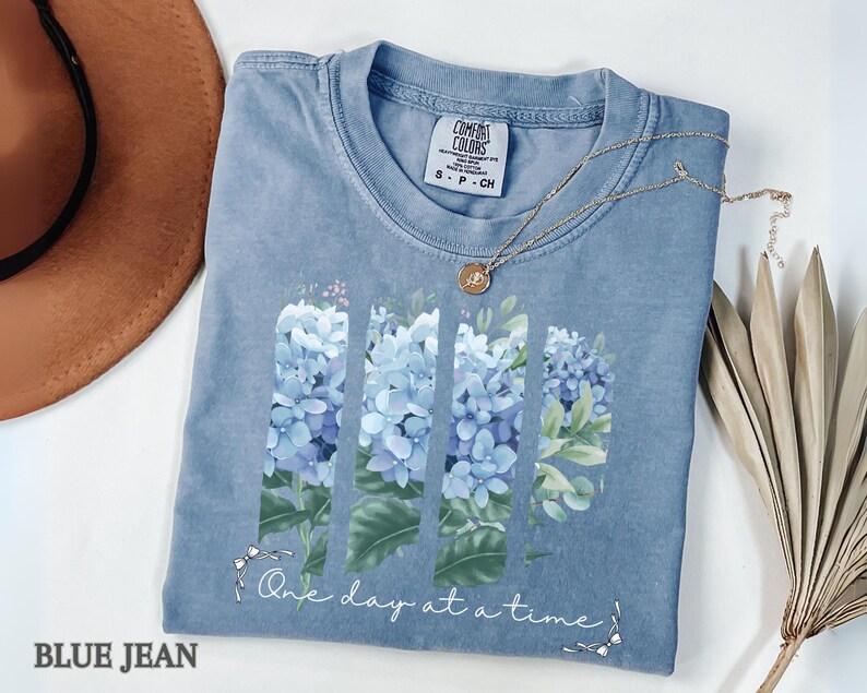 May include: A blue jean colored t-shirt featuring a graphic of four vertical panels filled with light blue hydrangea flowers and green leaves. Below the floral design, the text reads One day at a time in white cursive script with small bow accents.