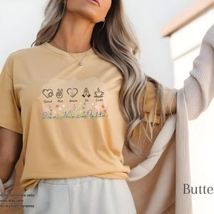 Puede incluir: Camiseta color mantequilla con un gr&aacute;fico floral y las palabras "Salud, Paz, Amor, Fe, Caf&eacute;" y "Que Nunca Falte". La camiseta tiene mangas cortas y cuello redondo.