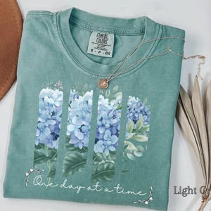 May include: A sage green short-sleeve t-shirt featuring a graphic of blue hydrangea flowers arranged in three vertical panels. Below the flowers, the text "One day at a time" is printed in white script. A silver necklace rests on the neckline.
