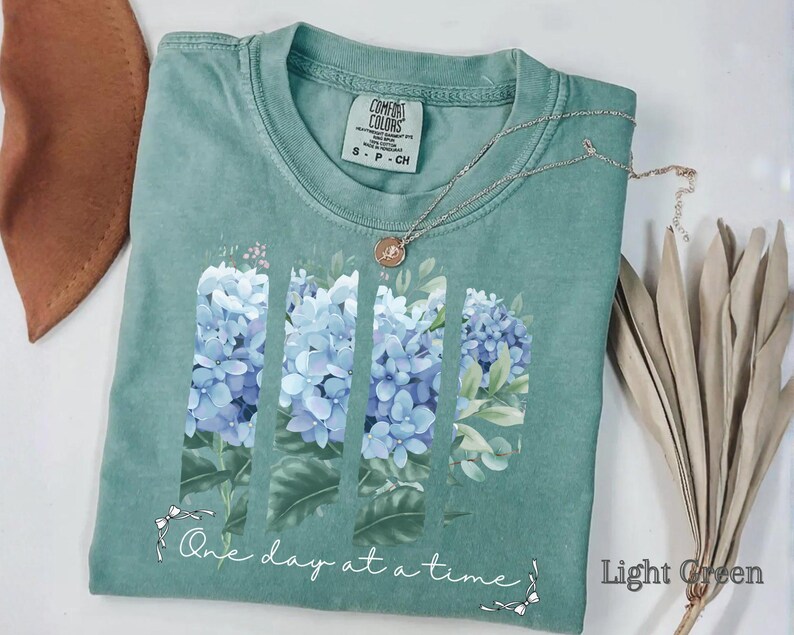 May include: A sage green short-sleeve t-shirt featuring a graphic of blue hydrangea flowers arranged in three vertical panels. Below the flowers, the text "One day at a time" is printed in white script. A silver necklace rests on the neckline.
