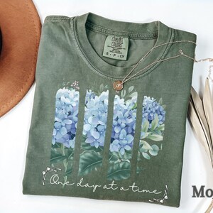 May include: A moss green t-shirt featuring a graphic of four vertical panels of blue hydrangea flowers and green leaves. Below the floral design, the text reads One day at a time in a white cursive script. The shirt has a crew neckline and a size S label.