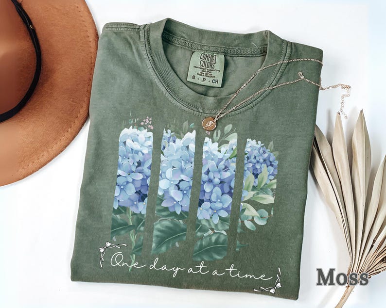 May include: A moss green t-shirt featuring a graphic of four vertical panels of blue hydrangea flowers and green leaves. Below the floral design, the text reads One day at a time in a white cursive script. The shirt has a crew neckline and a size S label.
