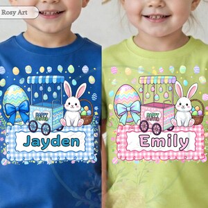 May include: Two children's t-shirts, one blue and one green, featuring Easter-themed designs. Each shirt has a cartoon bunny, an Easter egg, and a cart with the text "Happy Easter". The blue shirt says "Jayden" and the green shirt says "Emily".