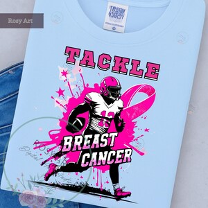 K&ouml;nnte beinhalten: Hellblaues T-Shirt mit rosa und wei&szlig;er Grafik. Die Grafik zeigt einen Footballspieler in rosa Helm und Uniform, ein rosa Band und die Worte "TACKLE BREAST CANCER".