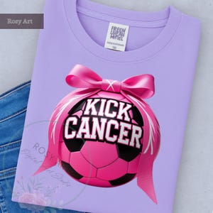 K&ouml;nnte beinhalten: Ein helllila Sweatshirt mit einer rosa Fu&szlig;ballgrafik. Der Fu&szlig;ball hat die Worte "KICK CANCER" in wei&szlig;er Schrift. Ein rosa Band ist um den Ball gebunden. Das Shirt ist auf einer blauen Jeans gefaltet.