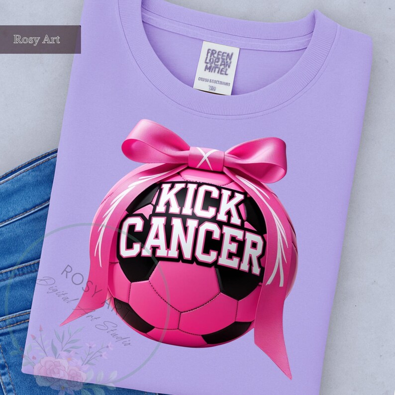 K&ouml;nnte beinhalten: Ein helllila Sweatshirt mit einer rosa Fu&szlig;ballgrafik. Der Fu&szlig;ball hat die Worte "KICK CANCER" in wei&szlig;er Schrift. Ein rosa Band ist um den Ball gebunden. Das Shirt ist auf einer blauen Jeans gefaltet.