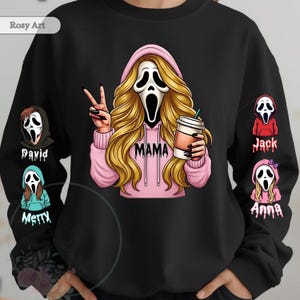 Puede incluir: Sudadera negra con una ilustración de dibujos animados de una máscara de Scream con pelo largo y rubio, con una sudadera con capucha rosa con la palabra "MAMA". El personaje sostiene una taza de café y hace el signo de la paz. Personajes adicionales de la máscara de Scream están en las mangas.