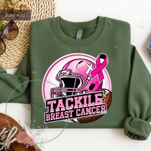 K&ouml;nnte beinhalten: Ein olivgr&uuml;nes Sweatshirt mit einem rosa Footballhelm, einem rosa Band und einem Football. Der Text "TACKLE BREAST CANCER" befindet sich darunter. Das Design ist auf der Vorderseite des Sweatshirts zentriert.