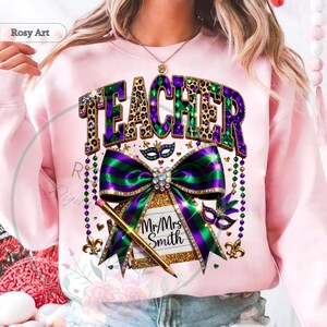 May include: A light pink sweatshirt with a graphic design featuring the word "TEACHER" in leopard print and purple and green colors. A large bow and pencil are also featured in the design, along with the text "Mr/Mrs Smith".