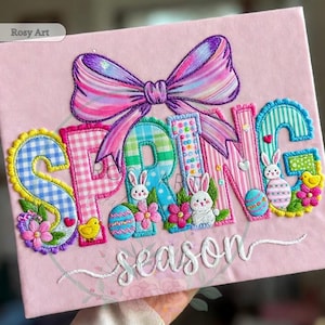 May include: A pink embroidered artwork with the word "SPRING" in colorful letters, decorated with Easter eggs, bunnies, and flowers. A large bow sits above the word, and the word "season" is embroidered below.