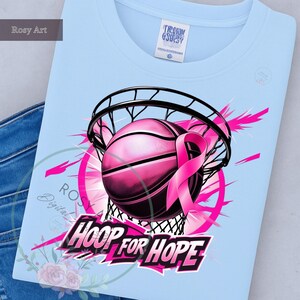 K&ouml;nnte beinhalten: Hellblaues T-Shirt mit einem pinkfarbenen Basketball, der durch ein Netz geht, einem pinkfarbenen Band und dem Schriftzug "HOOP FOR HOPE". Das Design umfasst pinkfarbene Akzente und ein florales Element. Das T-Shirt hat einen Rundhalsausschnitt.
