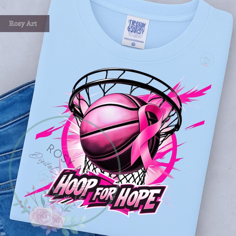 K&ouml;nnte beinhalten: Hellblaues T-Shirt mit einem pinkfarbenen Basketball, der durch ein Netz geht, einem pinkfarbenen Band und dem Schriftzug "HOOP FOR HOPE". Das Design umfasst pinkfarbene Akzente und ein florales Element. Das T-Shirt hat einen Rundhalsausschnitt.
