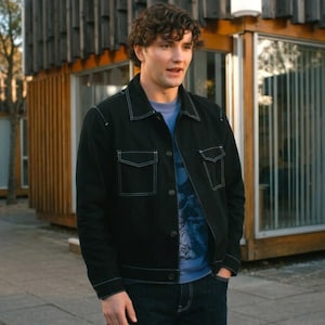 May include: A black denim jacket featuring prominent white contrast stitching, a classic collar, two button-flap chest pockets, and a button-front closure, worn over a light blue t-shirt with a dark graphic print and dark blue denim jeans.