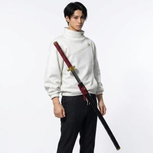 May include: A person wearing a white turtleneck sweatshirt and black trousers, holding a black and red katana sword. The sword has a gold hilt and a red handle wrap. The person is against a white background.