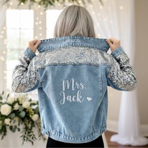 May include: Light blue denim jacket with silver sequin embellishments on the shoulders and sleeves. The back of the jacket has the text "Mrs. Jack" and a heart, with the date "07.12.25" above the collar.