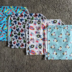 May include: Four drawstring bags with various patterns. One bag has race cars on a blue background, another features tractors on white, a third displays rockets on white, and the last shows astronauts on a light blue background.