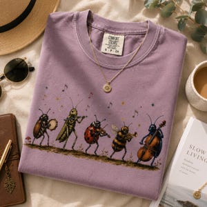 Könnte beinhalten: Ein malvenfarbenes T-Shirt mit Rundhalsausschnitt und einer Grafik von fünf Insekten, die Musikinstrumente spielen, darunter ein Käfer mit Tamburin, eine Heuschrecke mit Flöte, ein Marienkäfer mit Geige, eine Biene mit Trompete und ein blauer Käfer mit Cello.