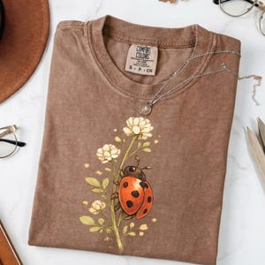 May include: A brown short-sleeved t-shirt featuring a graphic of a red ladybird climbing a stem with pale yellow flowers. The shirt has a Comfort Colors brand label at the inner neckline with the text S P CH printed on the tag.