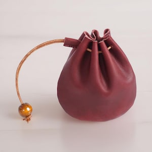 May include: A burgundy leather drawstring pouch with a brown leather cord and a wooden bead. The pouch is closed and has a round shape.