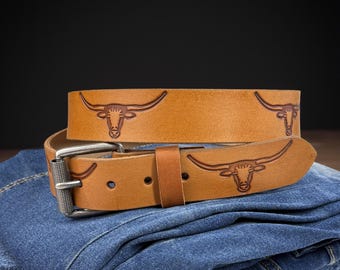 May include: A brown leather belt with a silver buckle and an embossed design of a longhorn steer. The belt is folded and placed on a pair of blue jeans.