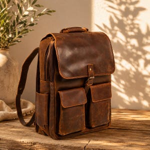 May include: A brown leather backpack with a flap closure and two front pockets. The backpack has adjustable shoulder straps and a top handle. The leather has a distressed look, and the bag appears to be new.