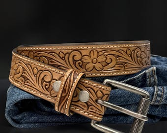 May include: A light brown leather belt with a floral embossed design. The belt features a silver-colored buckle and two silver rivets. The belt is resting on a pair of blue jeans.