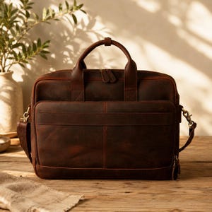 Puede incluir: Un maletín de cuero marrón con asa superior y bandolera desmontable. La bolsa tiene múltiples compartimentos, incluyendo un bolsillo frontal y un compartimento principal con cremallera. El cuero tiene un aspecto envejecido con costuras rojas.