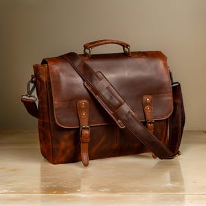 May include: A brown leather messenger bag with a long adjustable strap and two buckles on the front. The bag has a distressed look and is made of high-quality leather.