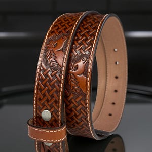 May include: A brown leather belt with a woven pattern and embossed deer heads. The belt has a silver buckle and a matching leather loop. The stitching is white.
