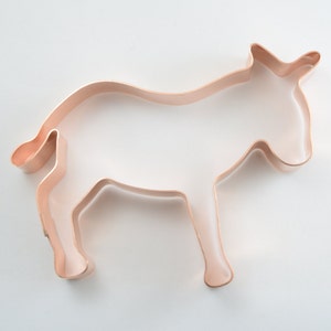 May include: A copper-colored metal cookie cutter shaped like a donkey. The outline features the distinct silhouette of the animal, including the ears, head, body, four legs, and a small tail, with a smooth, metallic finish and open center.