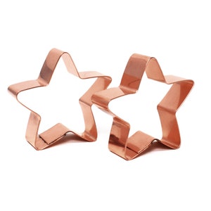 May include: Two copper star-shaped cookie cutters. The cutters are slightly different sizes and are lying on a white surface.