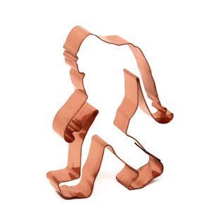 Pode incluir: Um cortador de biscoitos de cobre na forma de um Pé Grande caminhando. O cortador é detalhado com uma cabeça arredondada, braços, pernas e pés.