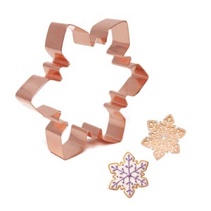 May include: A copper snowflake cookie cutter with two decorated snowflake biscuits. One biscuit is decorated with gold icing and the other with white icing and purple details.