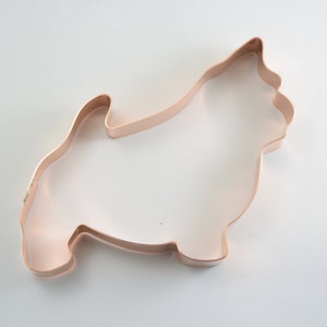 May include: A copper-colored metal cookie cutter shaped like a small dog with pointed ears and a short, upright tail. The outline of the dog is clearly defined by the thin, continuous metal strip that forms the frame of this baking tool.