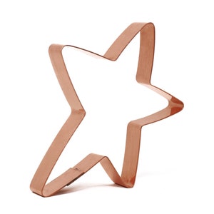 May include: A copper-colored star-shaped cookie cutter. The cutter is made of a single piece of metal bent into the shape of a five-pointed star. The star is tilted slightly to the side, giving it a dynamic appearance.