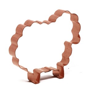 May include: A copper-coloured cookie cutter shaped like a sheep. The cutter has a wavy outline and a flat bottom.