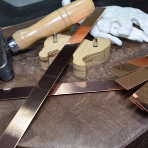 May include: A close-up of a wooden workbench with a variety of tools and materials, including a wooden mallet, a metal scraper, and several strips of copper metal.