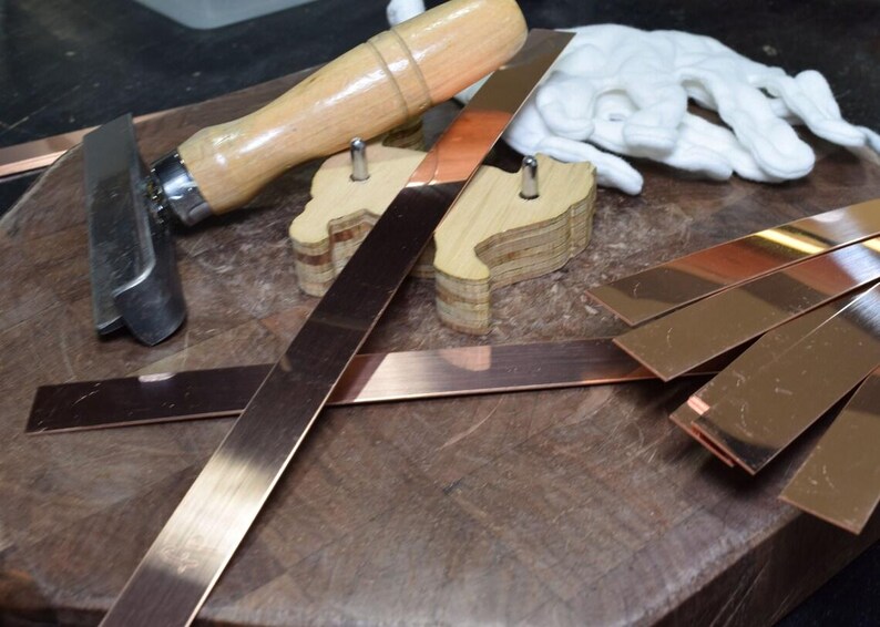 May include: A close-up of a wooden workbench with a variety of tools and materials, including a wooden mallet, a metal scraper, and several strips of copper metal.