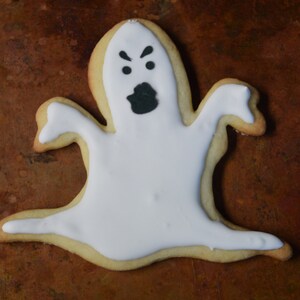 May include: A white ghost-shaped sugar cookie with black icing for the eyes and mouth. The cookie is on a brown surface.