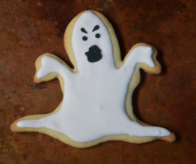 May include: A white ghost-shaped sugar cookie with black icing for the eyes and mouth. The cookie is on a brown surface.