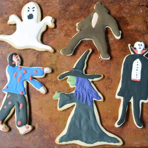May include: Five Halloween-themed sugar cookies decorated with icing. The cookies are shaped like a ghost, a zombie, a witch, a vampire, and a monster. The cookies are on a brown metal baking sheet.