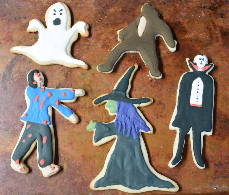 May include: Five Halloween-themed sugar cookies decorated with icing. The cookies are shaped like a ghost, a zombie, a witch, a vampire, and a monster. The cookies are on a brown metal baking sheet.