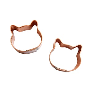May include: Two copper-colored cat-shaped cookie cutters. Each cutter has a simple outline of a cat's head with pointed ears and a rounded face. The cutters are made of thin metal and have a slight sheen.