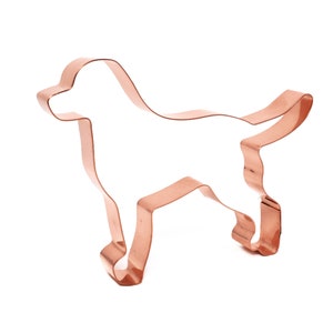 May include: A copper-coloured dog-shaped cookie cutter. The cutter is a continuous line, depicting a dog in a standing position with its tail up. The cutter is made of copper-coloured metal and is set against a white background.