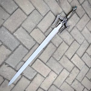 May include: A long, silver sword with an ornate handle and guard. The blade is straight and tapers to a point. The handle features detailed carvings and a green gem at the pommel. The sword is lying on a brick surface.