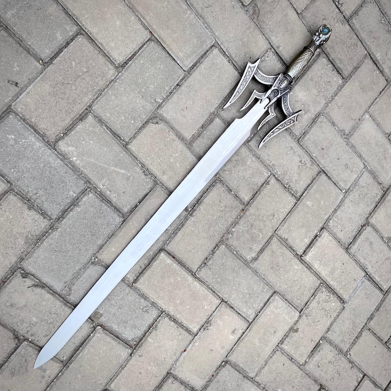 May include: A long, silver sword with an ornate handle and guard. The blade is straight and tapers to a point. The handle features detailed carvings and a green gem at the pommel. The sword is lying on a brick surface.