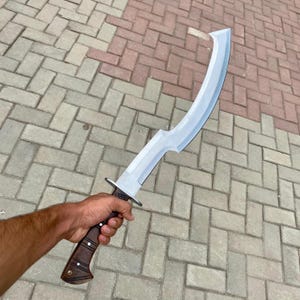 May include: A silver curved sword with a brown wooden handle. The sword is held by a hand in the image.