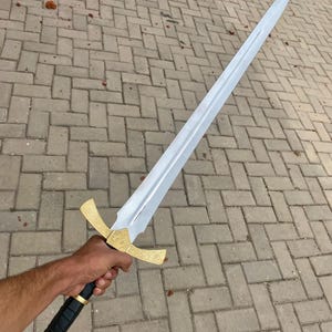 May include: A silver sword with a gold hilt and a red jewel on the pommel. The sword is held by a hand against a background of gray brick.
